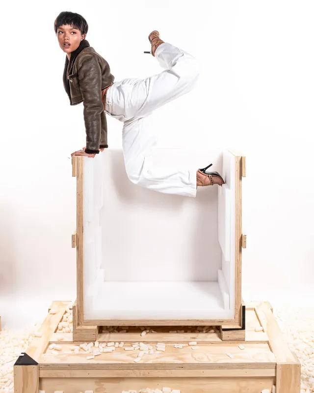 Whitney Peak posing on a studio platform in white trousers, brown jacket and heels.