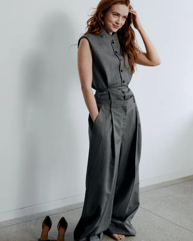 Sadie Sink standing barefoot beside removed black heels in a grey outfit.
