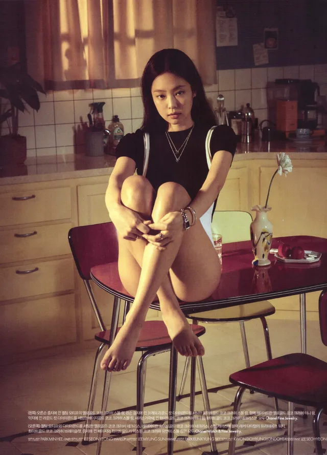 Jennie Kim sitting barefoot on a stool in a dark outfit during an indoor portrait.