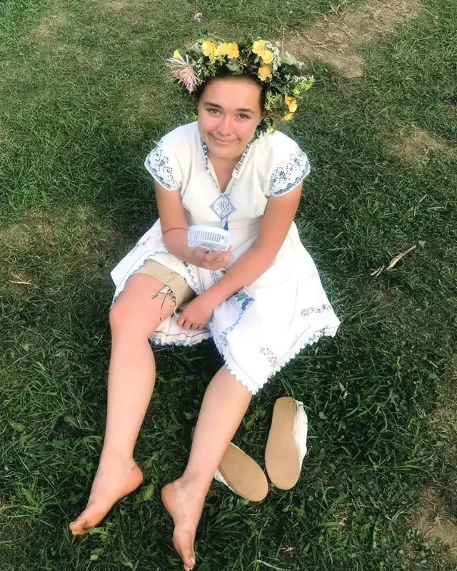 Florence Pugh sitting barefoot on grass in a white dress and flower crown.