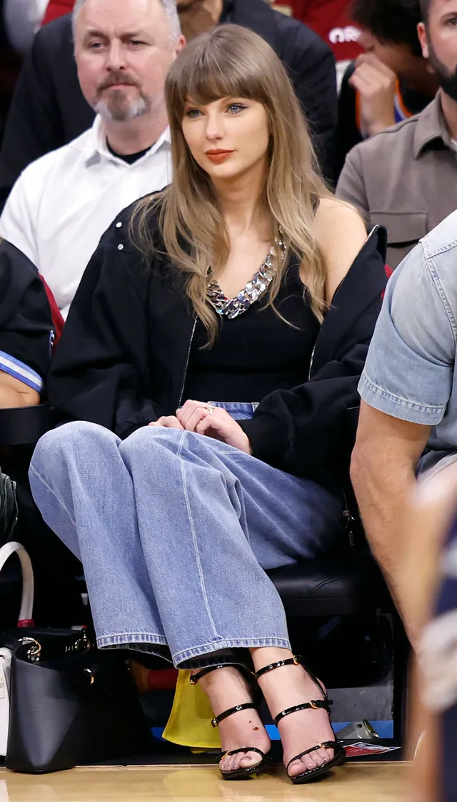 Taylor Swift: seated at an event in blue trousers and black heels.