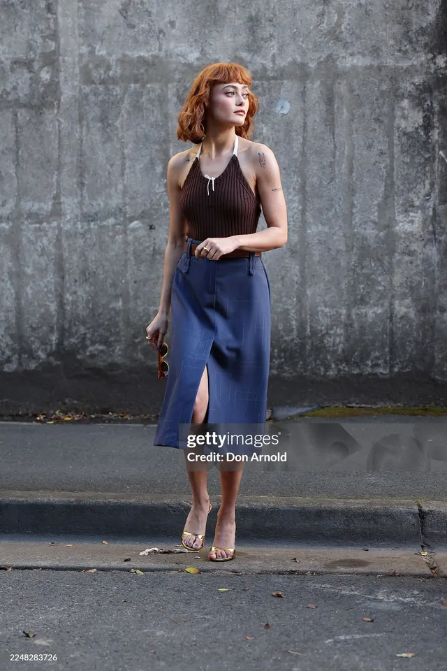 Ella Purnell standing outside in a blue skirt and pale sandals.