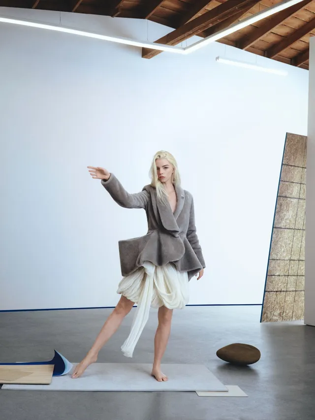 Anya Taylor-Joy posing barefoot in a skirt and gray jacket.
