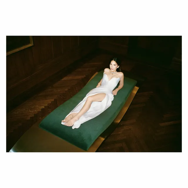 Olivia Munn reclining barefoot in a white dress on a green bench.