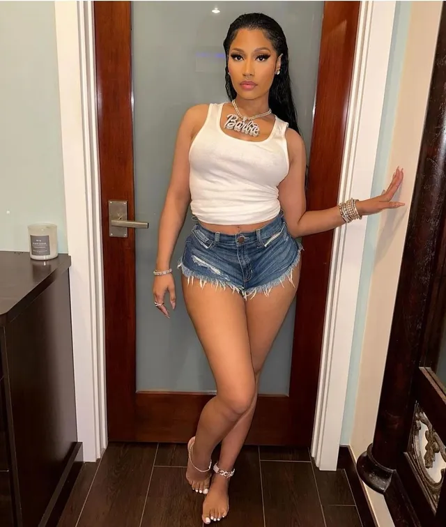 Nicki Minaj standing barefoot in a doorway in a white top and denim shorts.