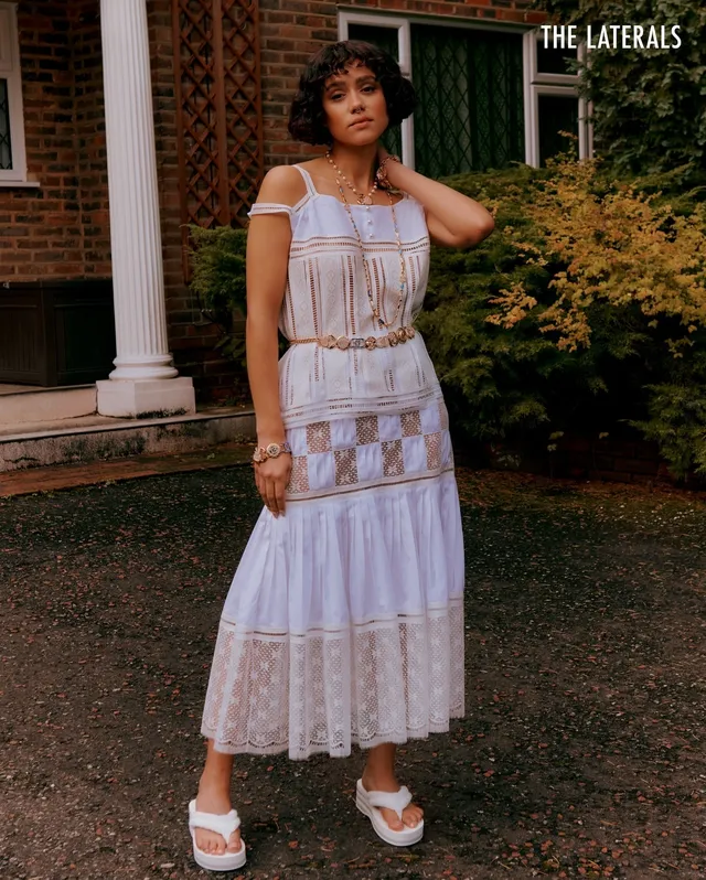Nathalie Emmanuel standing outdoors in a white dress and white sandals.