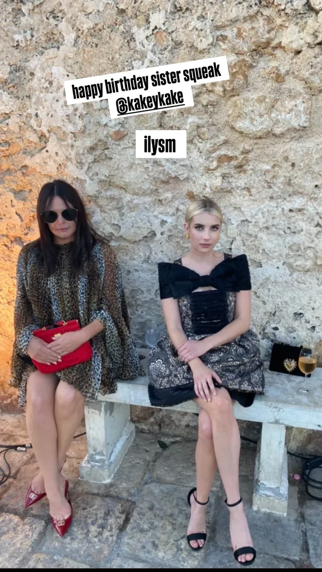 Emma Roberts sitting outdoors in a black dress and strappy heels.