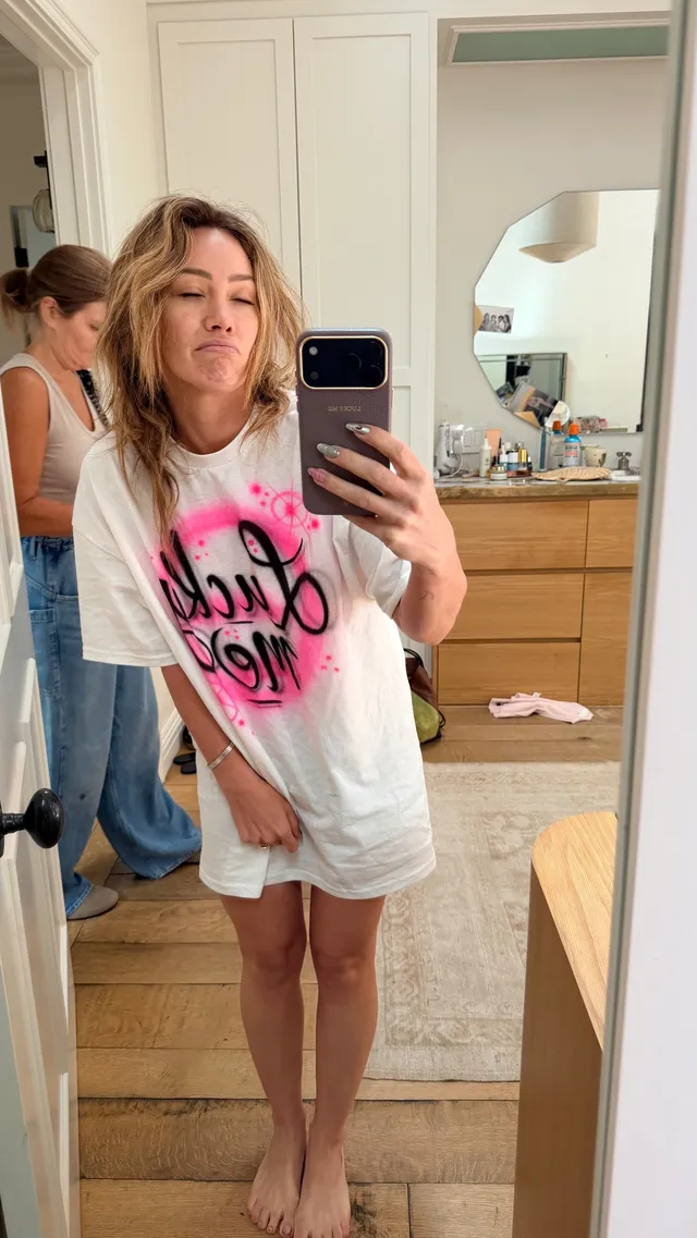 Hilary Duff taking a barefoot mirror photo in a white T-shirt.