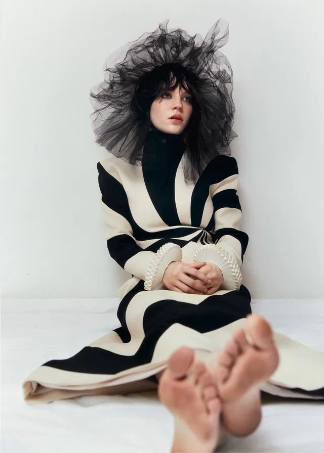 Sophie Thatcher sitting on a white floor with her bare soles facing the camera.