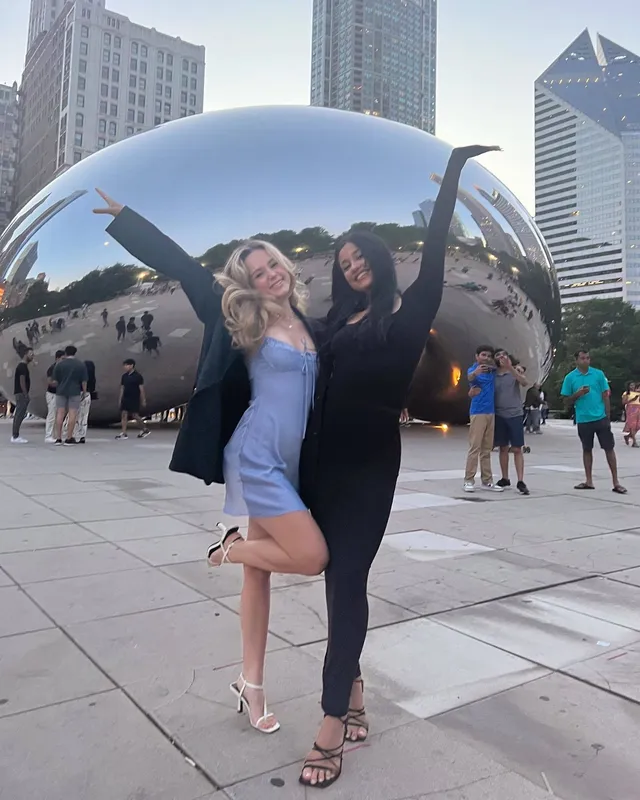 Brec Bassinger posing with a friend in a pale blue dress and white heels by a city sculpture.