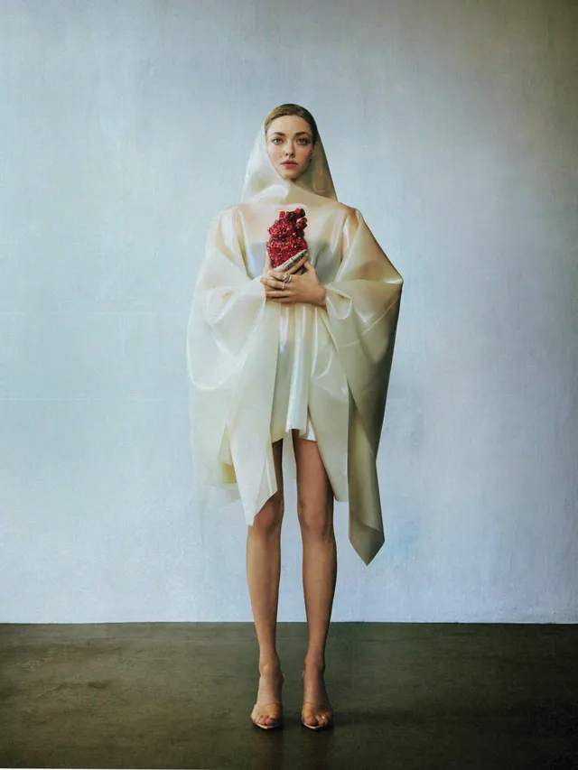 Amanda Seyfried standing in a white outfit and heels holding flowers.
