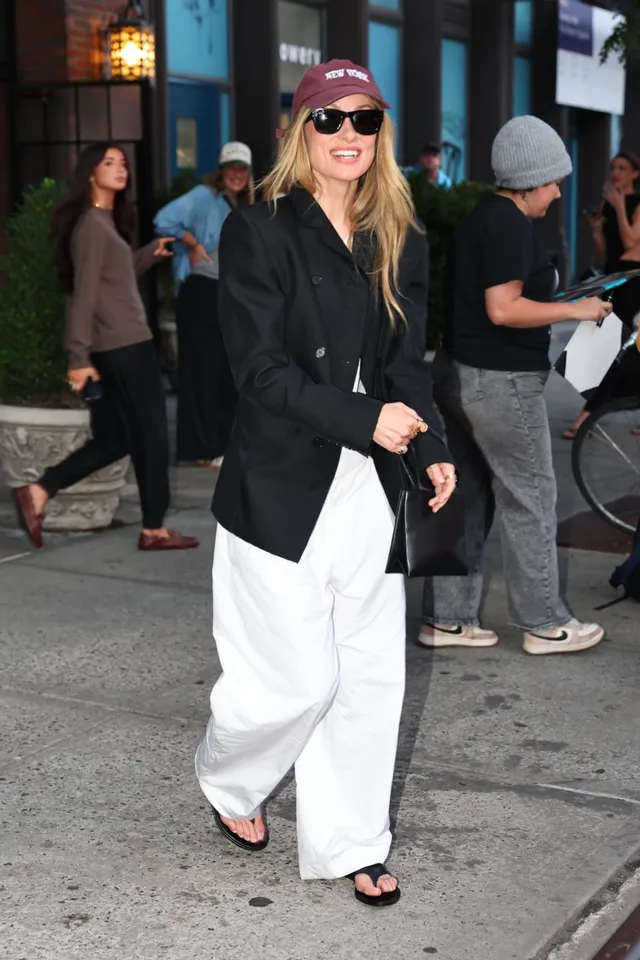 Olivia Wilde walking along a street in white trousers and flip-flops.