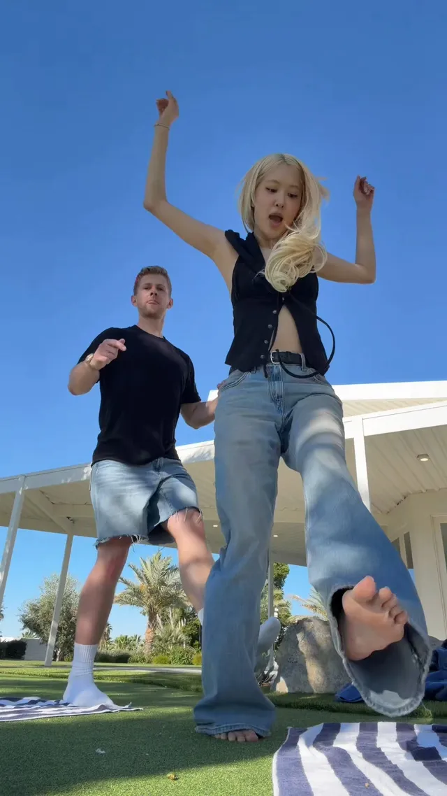 Rosé lifting a bare foot toward the camera while dancing outside in blue jeans.