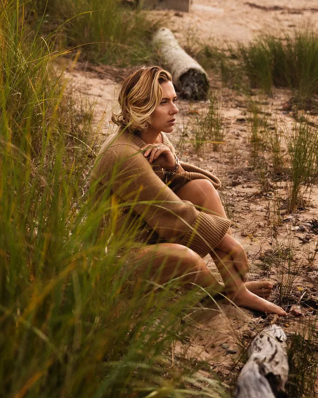 Scarlett Johansson: seated barefoot on a beach beside tall grass.