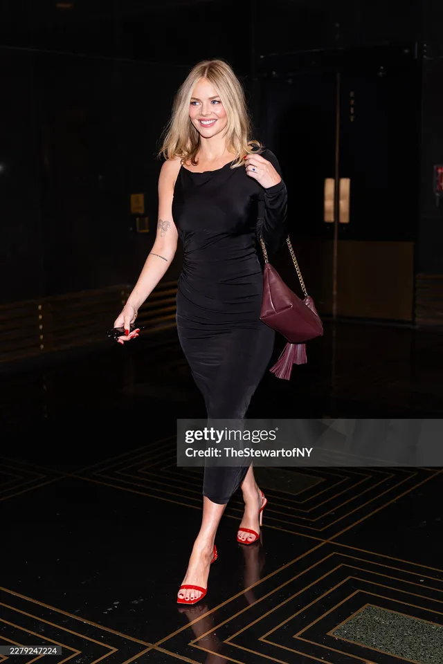 Samara Weaving posing in a black dress and red heels indoors.