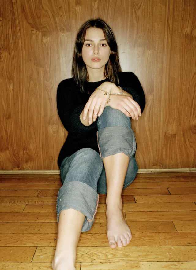 Keira Knightley seated on a wooden floor in a black top and jeans.