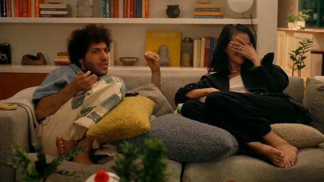 Selena Gomez sitting barefoot beside a man on a living room sofa.