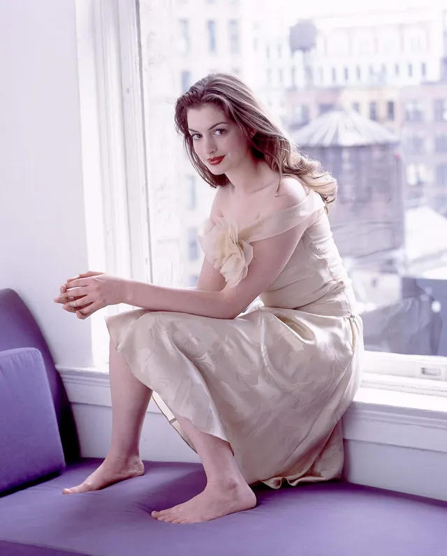 Anne Hathaway seated barefoot on a purple bench beside a window.