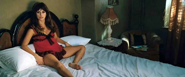 Penélope Cruz sitting barefoot on a bed in a red outfit.