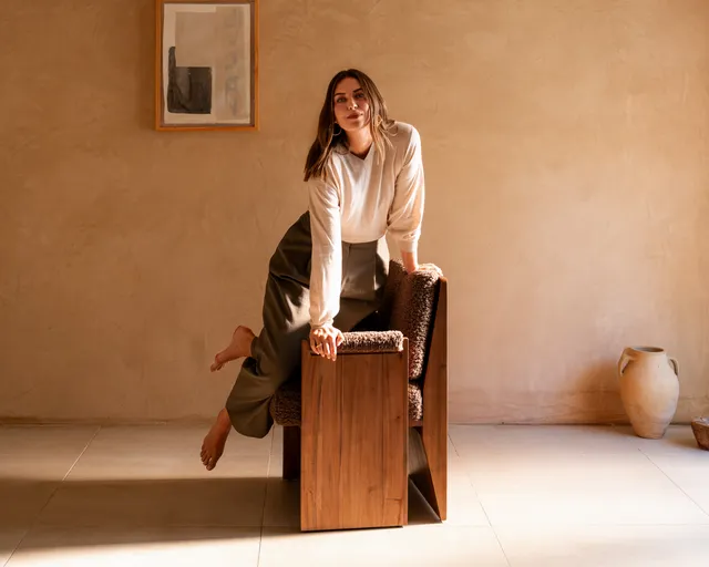 Maria Sharapova sitting barefoot on a wooden stool in a pale blouse and dark trousers.