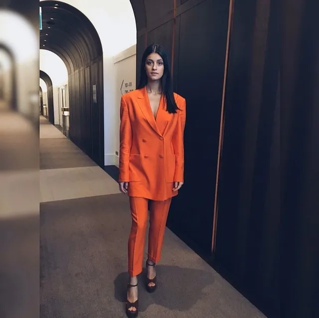 Anya Chalotra wearing an orange suit and black heels in a corridor.