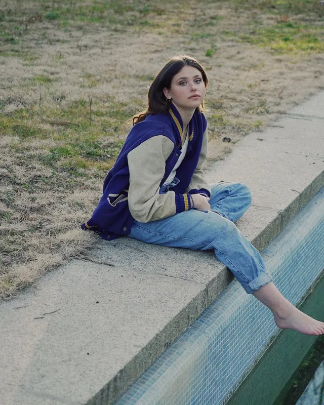 Nicole Wallace sitting beside a pool in jeans and a varsity jacket with a bare foot in the water.