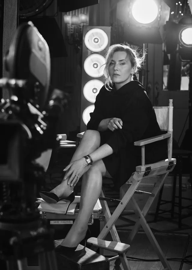Kate Winslet sitting indoors in a dark outfit and boots.