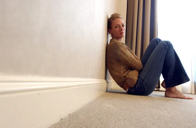 Uma Thurman sitting barefoot against a wall in jeans.