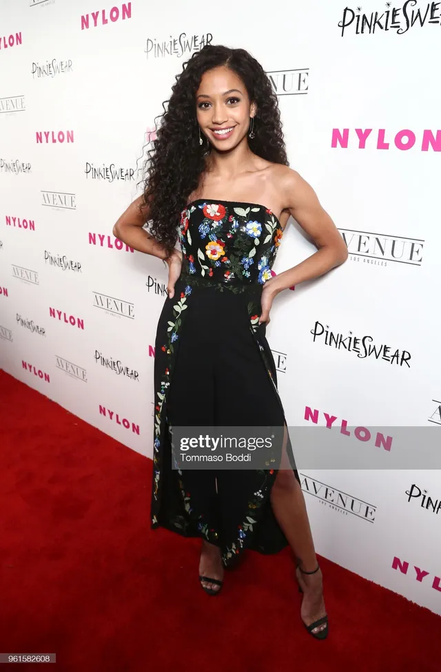 Samantha Logan posing in a black floral dress and strappy heels on a carpet.