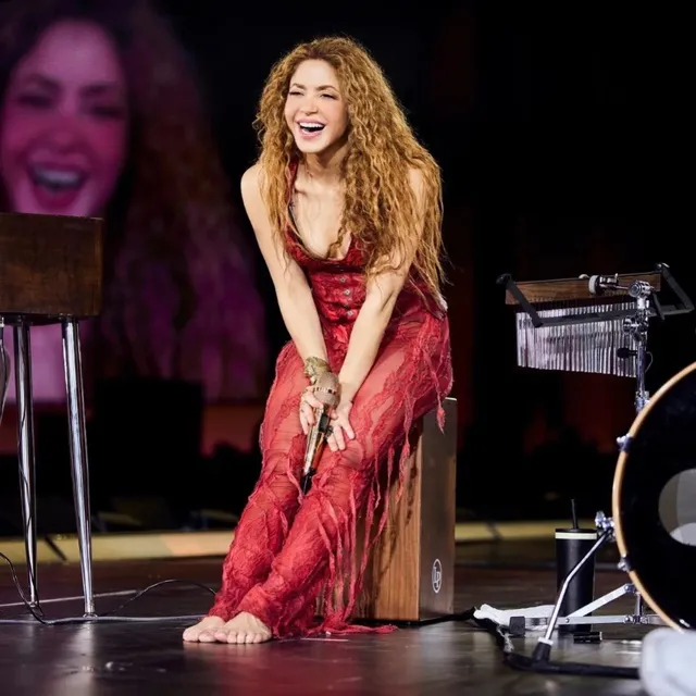 Shakira kneeling barefoot during a stage performance in a red dress.