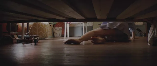 Chloë Grace Moretz with bare feet visible beneath a bed in a screen image.