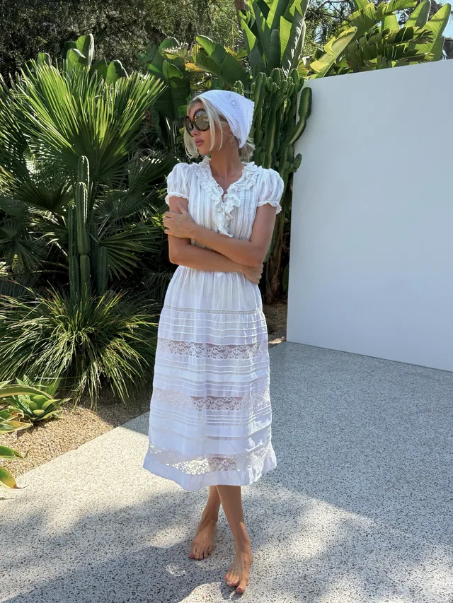 Elsa Hosk standing barefoot beside tropical plants in a white dress.