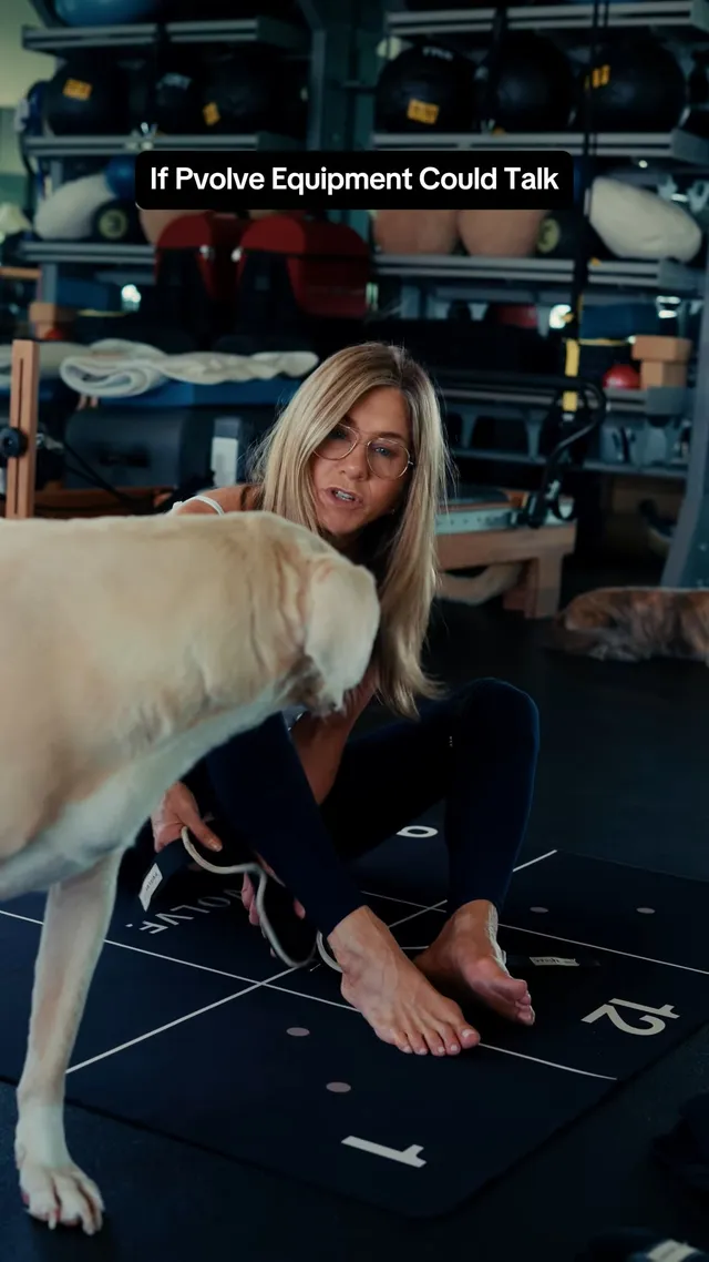 Jennifer Aniston sitting barefoot on an exercise mat beside a dog.