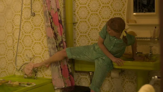 Gemma Arterton standing in a patterned room in a green dress in a screen scene.