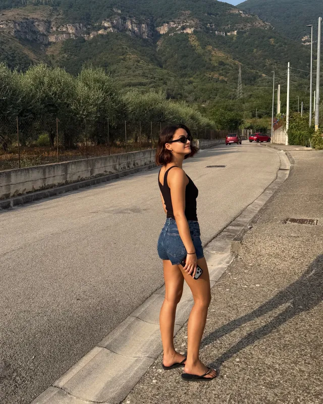 Camila Mendes standing outdoors in denim shorts and sandals.