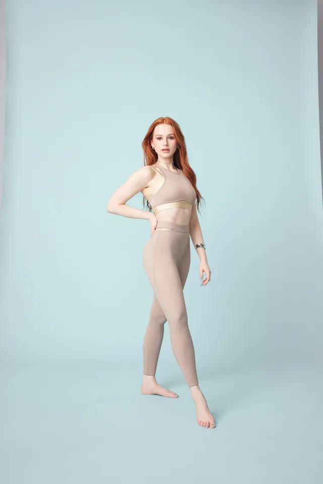 Madelaine Petsch posing barefoot in pale activewear against a blue backdrop.
