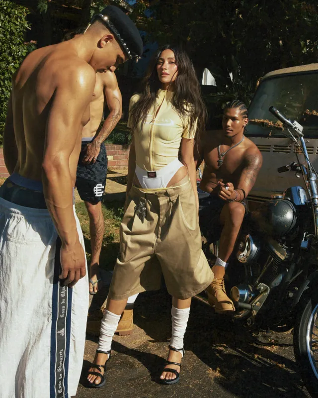 Shay Mitchell posing beside a motorbike in cargo trousers and sandals.