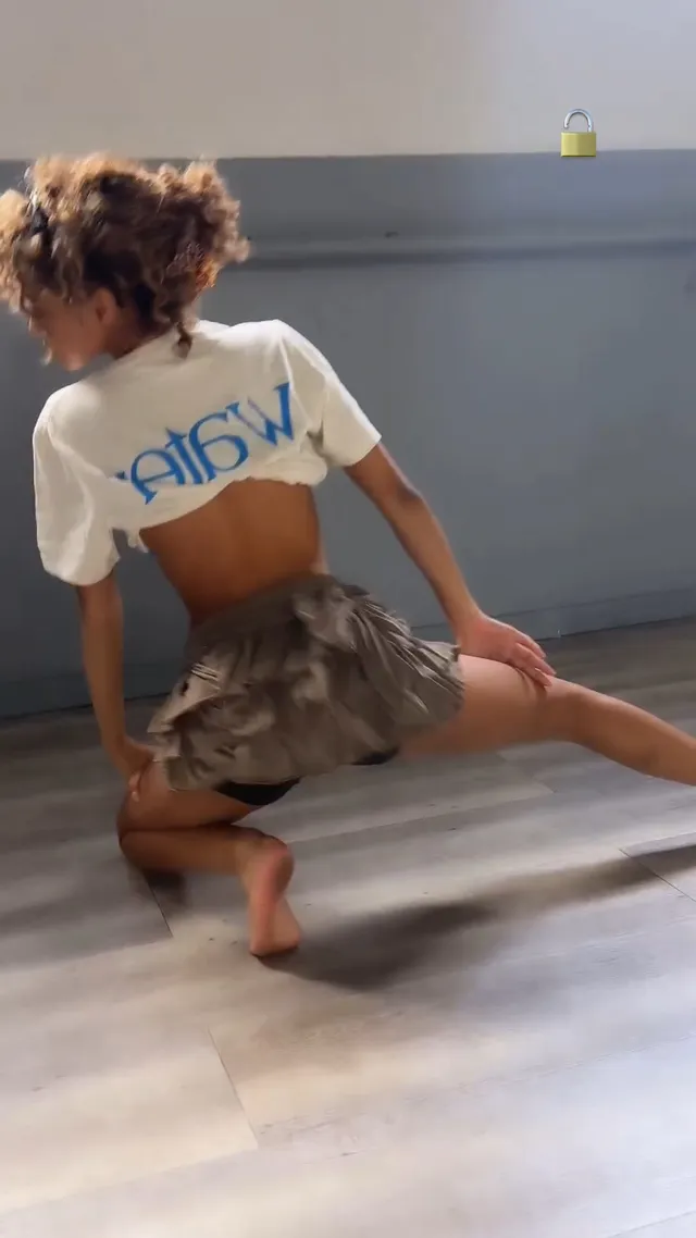 Tyla stretching barefoot on a wooden floor in a short patterned skirt.