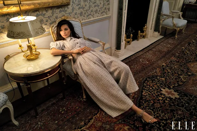 Gemma Chan reclining in a hotel chair in a long dress.
