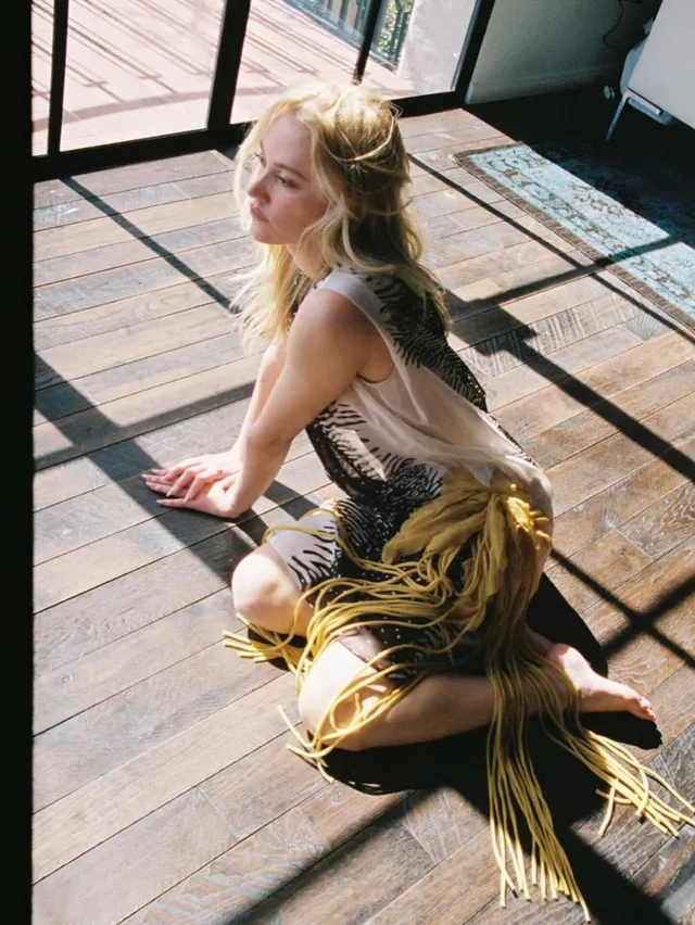 Maika Monroe sitting barefoot beside large windows.