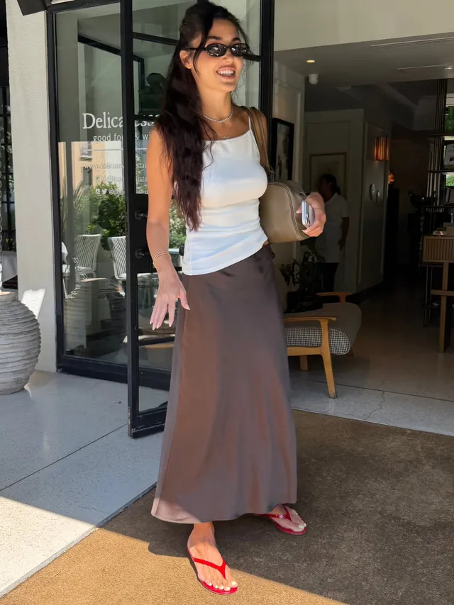 Hande Erçel standing outside a shop in a white top, long grey skirt and red flip-flops.