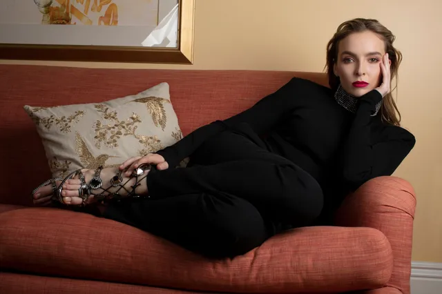 Jodie Comer reclining on a sofa in a black outfit and sandals.