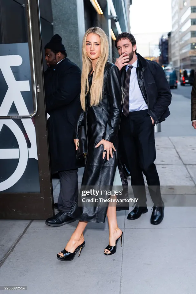 Grace Van Patten standing outdoors in a black coat and black heels.