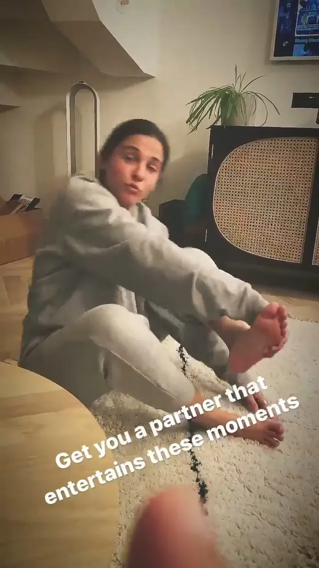 Naomi Scott sitting in a gray outfit with a bare sole extended.