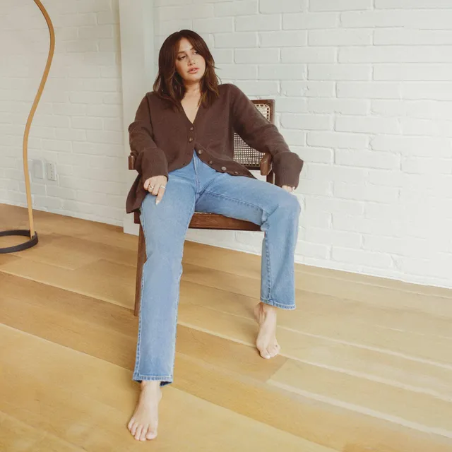 Ashley Tisdale sitting barefoot in a chair wearing jeans and a brown cardigan.