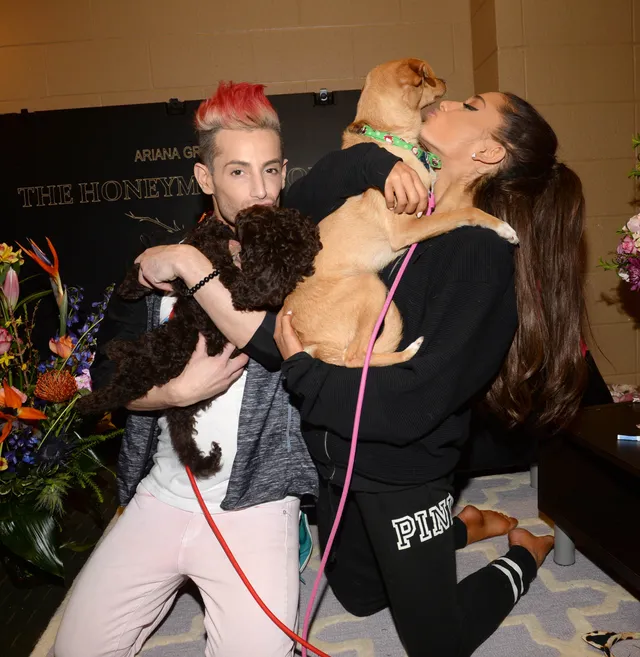 Ariana Grande kneeling barefoot on a bed while holding a dog beside another guest.