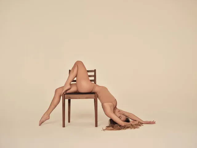 Sydney Sweeney reclining across a wooden chair against a cream backdrop.