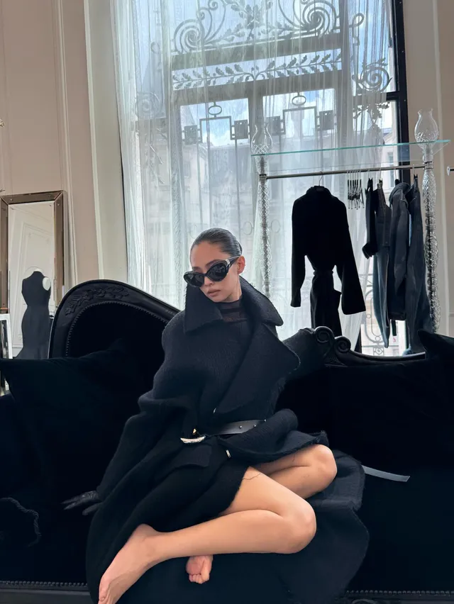 Jennie Kim sitting barefoot in a dark coat on an ornate chair.
