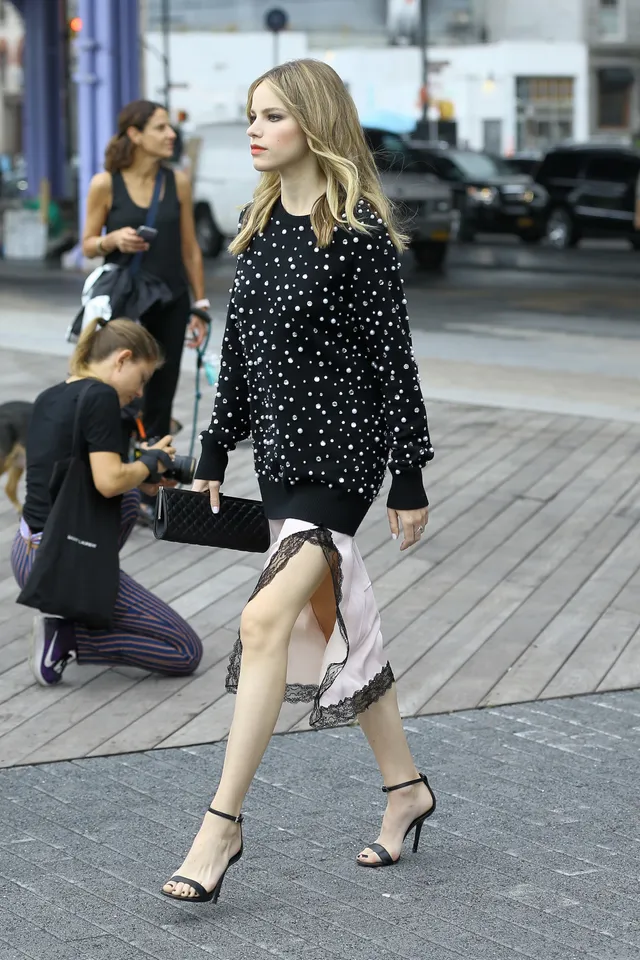 Halston Sage walking outdoors in a patterned skirt, dark jacket and black heels.