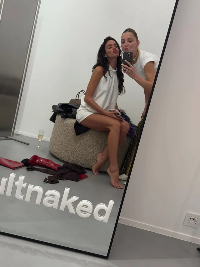 Bruna Marquezine taking a mirror selfie with a friend while sitting barefoot.